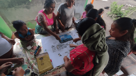 A group of women and children stand around a table on which they are using pictures and words to create a story of their village..
